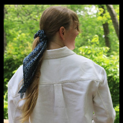 A woman viewed from behind wearing a white shirt and a dark blue patterned hair scarf tied in her ponytail, set against a lush green outdoor background.