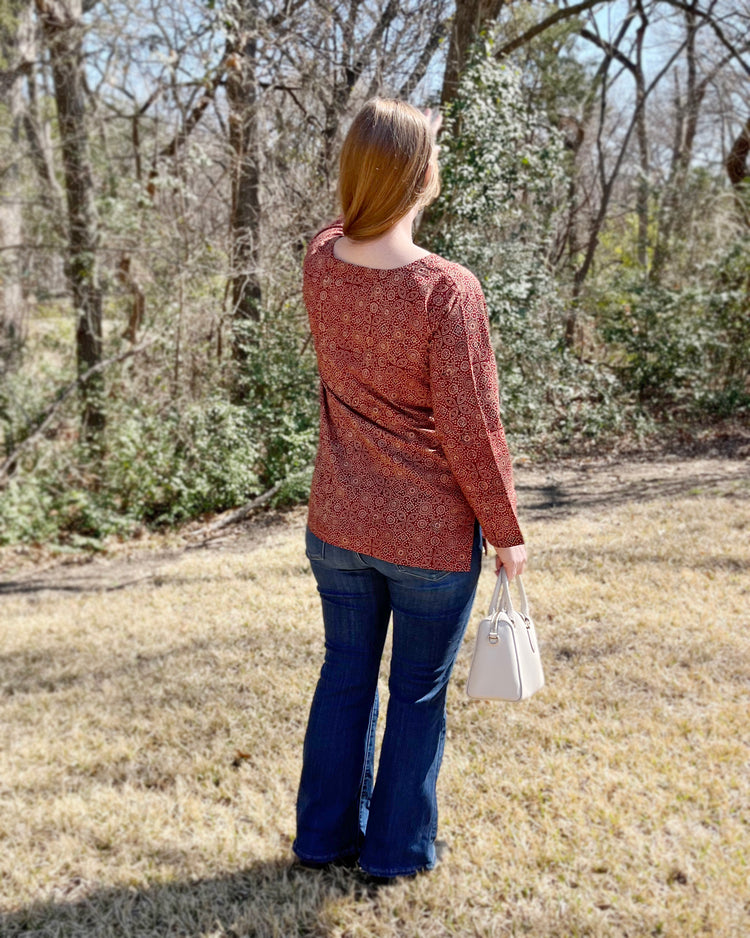A person standing in a natural outdoor setting wearing a red ajrak shirt with mirror work, full sleeves, and a round neckline.