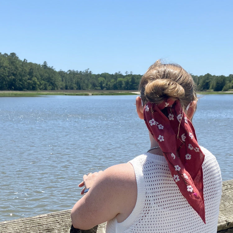 A woman seen from behind wearing a red hand-embroidered bandana with a white cherry blossom pattern tied in her hair, standing on a wooden dock overlooking a calm lake under a clear blue sky.